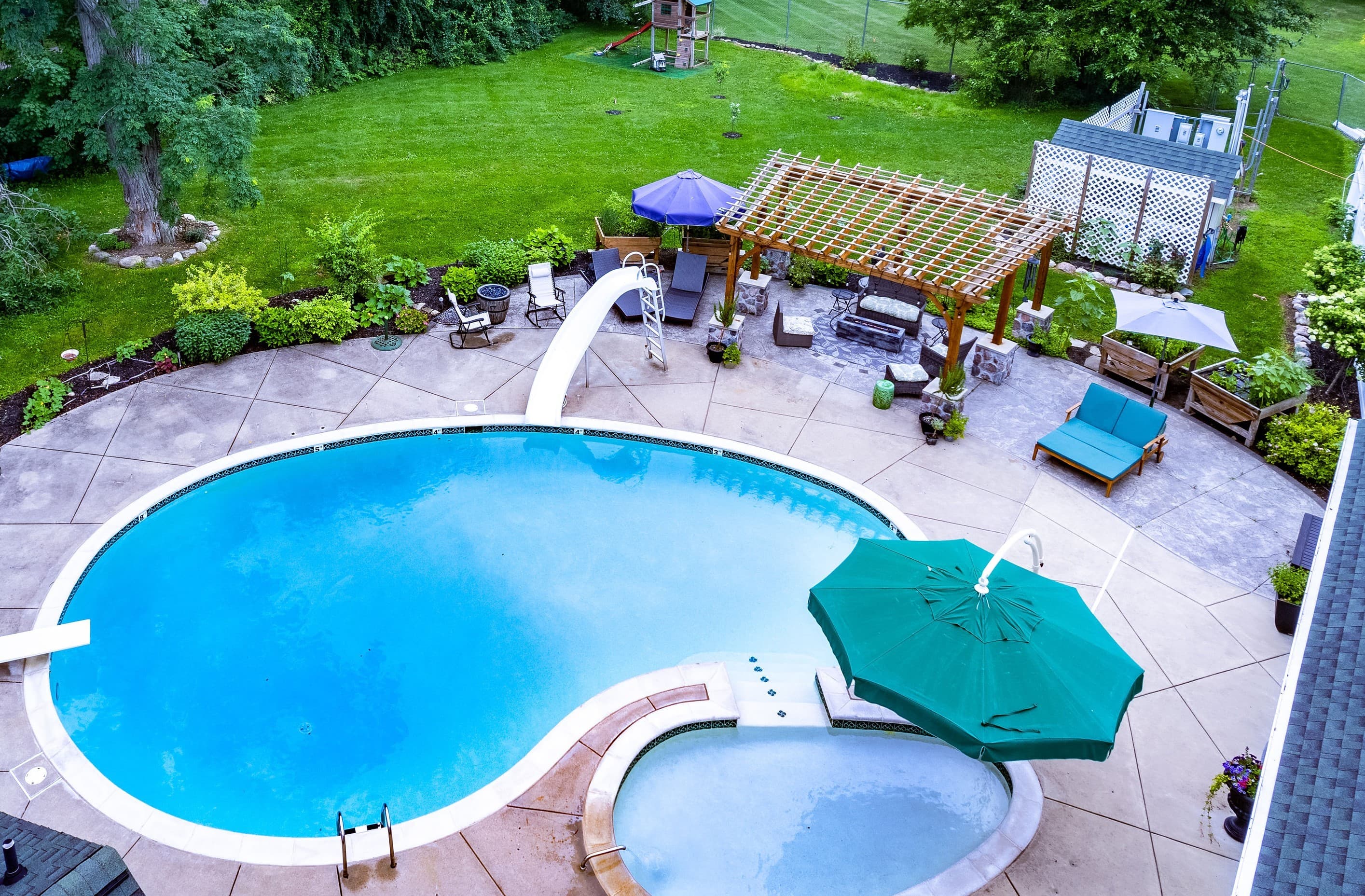 Aerial view of the outdoor pool area at Maple Cove Bed & Breakfast featuring a kidney-shaped swimming pool with a slide, a hot tub, and a furnished patio with a wooden pergola.