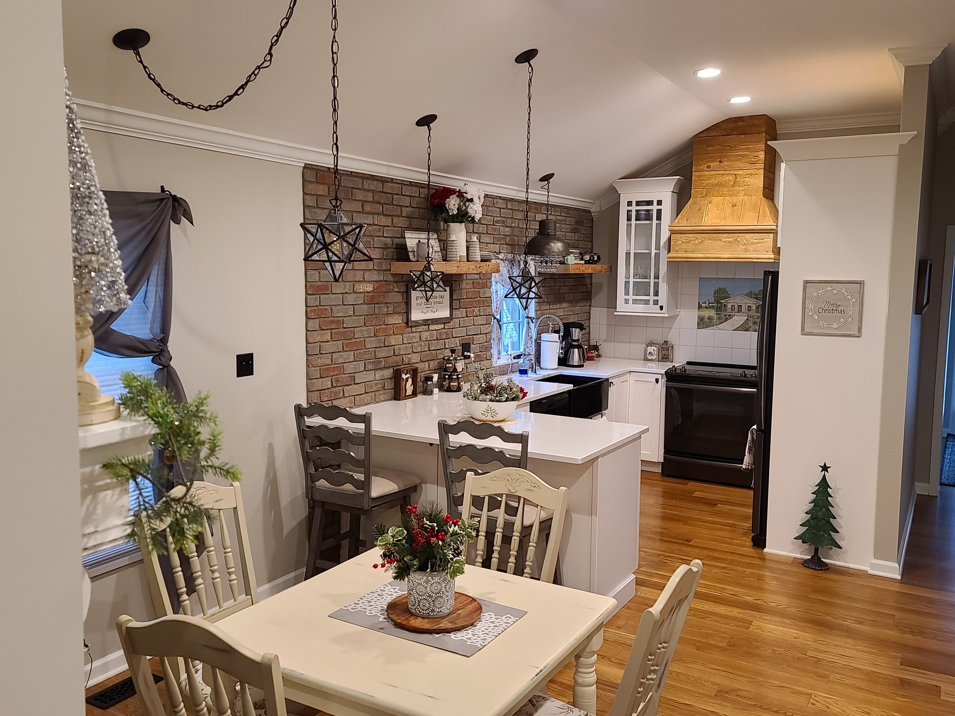 Farmhouse kitchen and dining area featuring a brick accent wall, white cabinetry with black appliances, a custom wooden range hood, and a dining table adorned with festive winter decor.