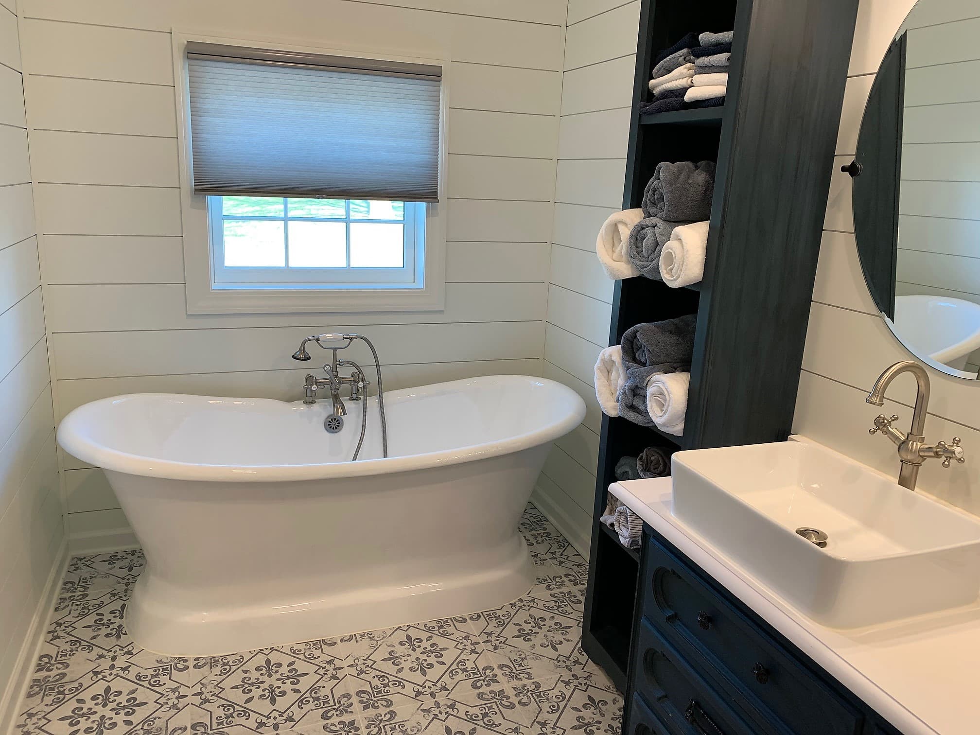 Farmhouse bathroom at The August House featuring a white freestanding soaking tub under a window, white shiplap walls, a dark storage tower with towels, and a rectangular vessel sink.