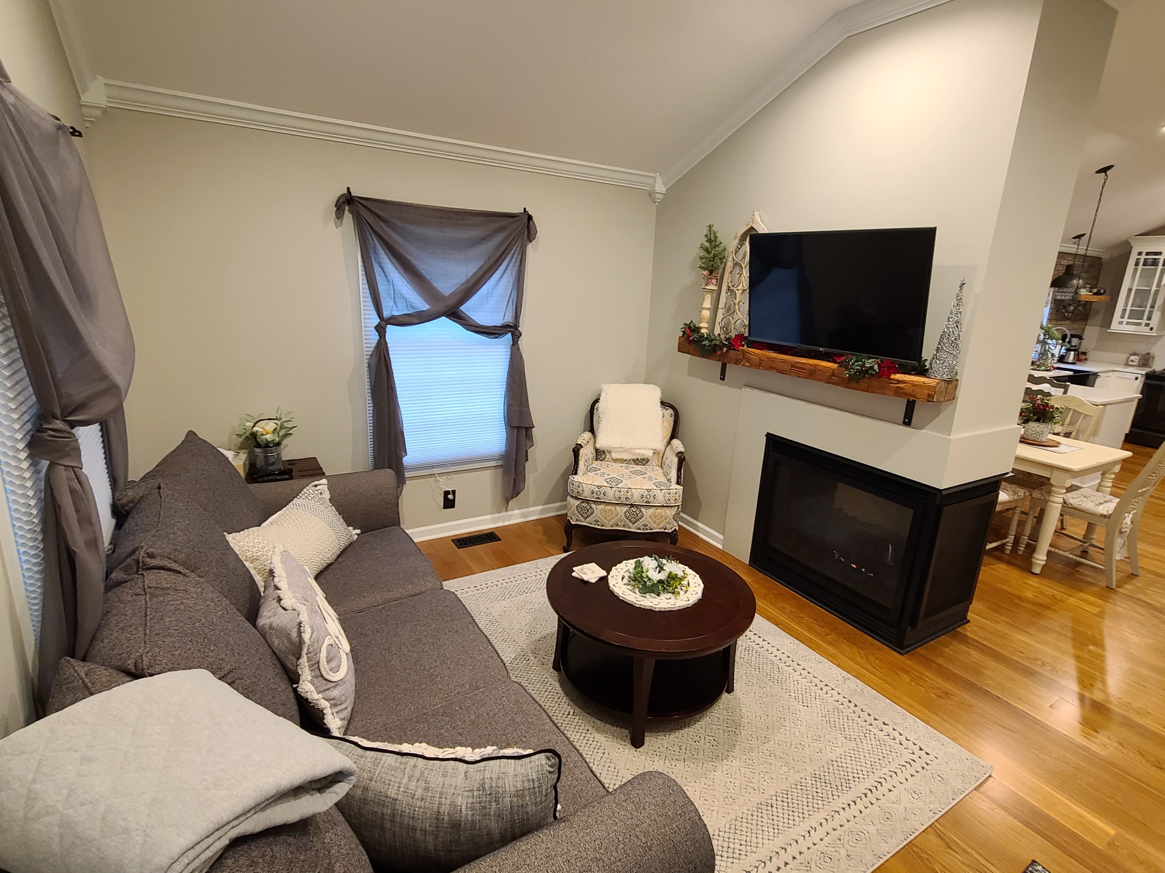 Living room area at The August House featuring a grey sofa, patterned armchair, corner fireplace with a mounted TV, and hardwood floors.