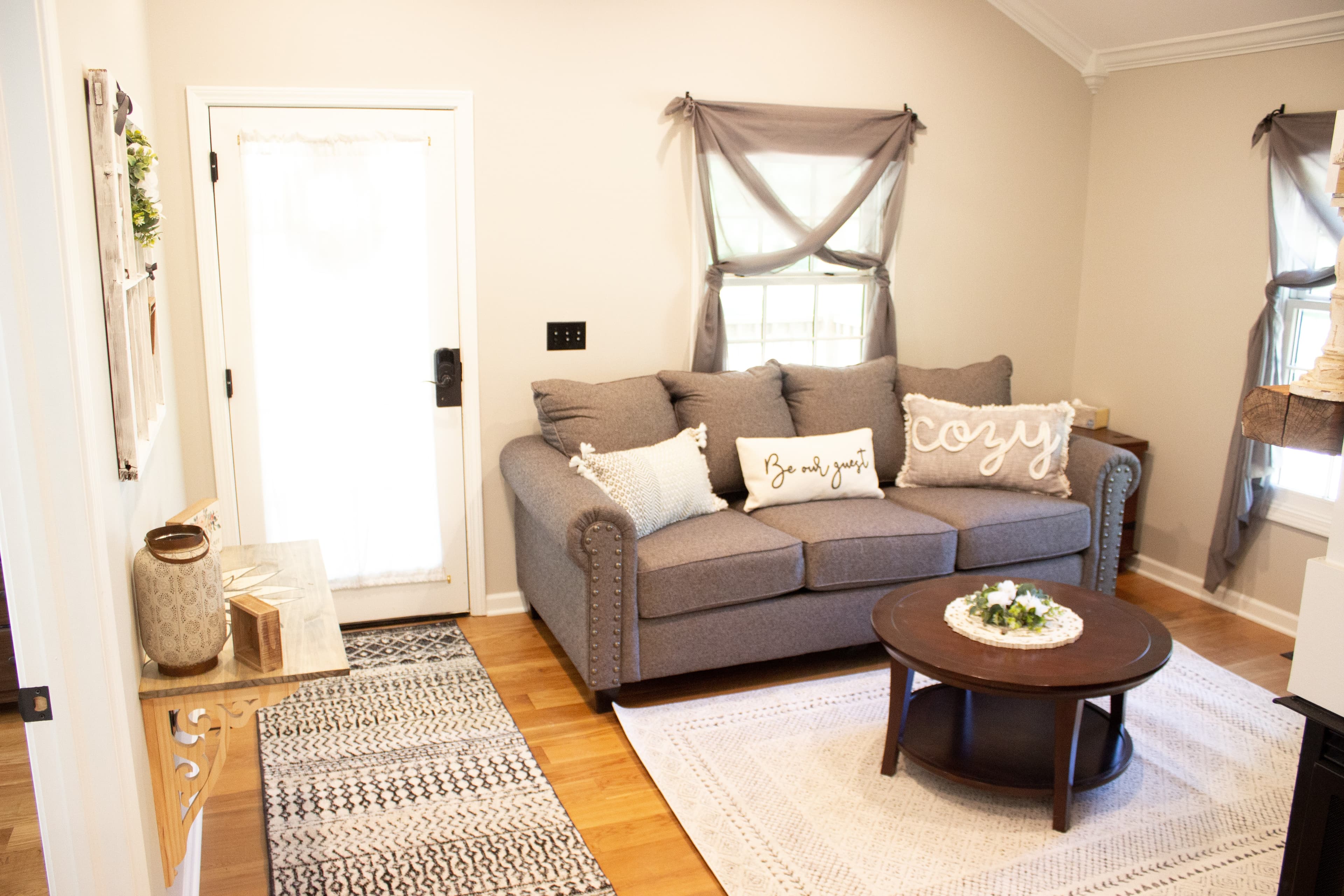 Living space at The August House featuring a grey sofa with 'Cozy' and 'Be our guest' throw pillows, a round wooden coffee table, and a white front door.