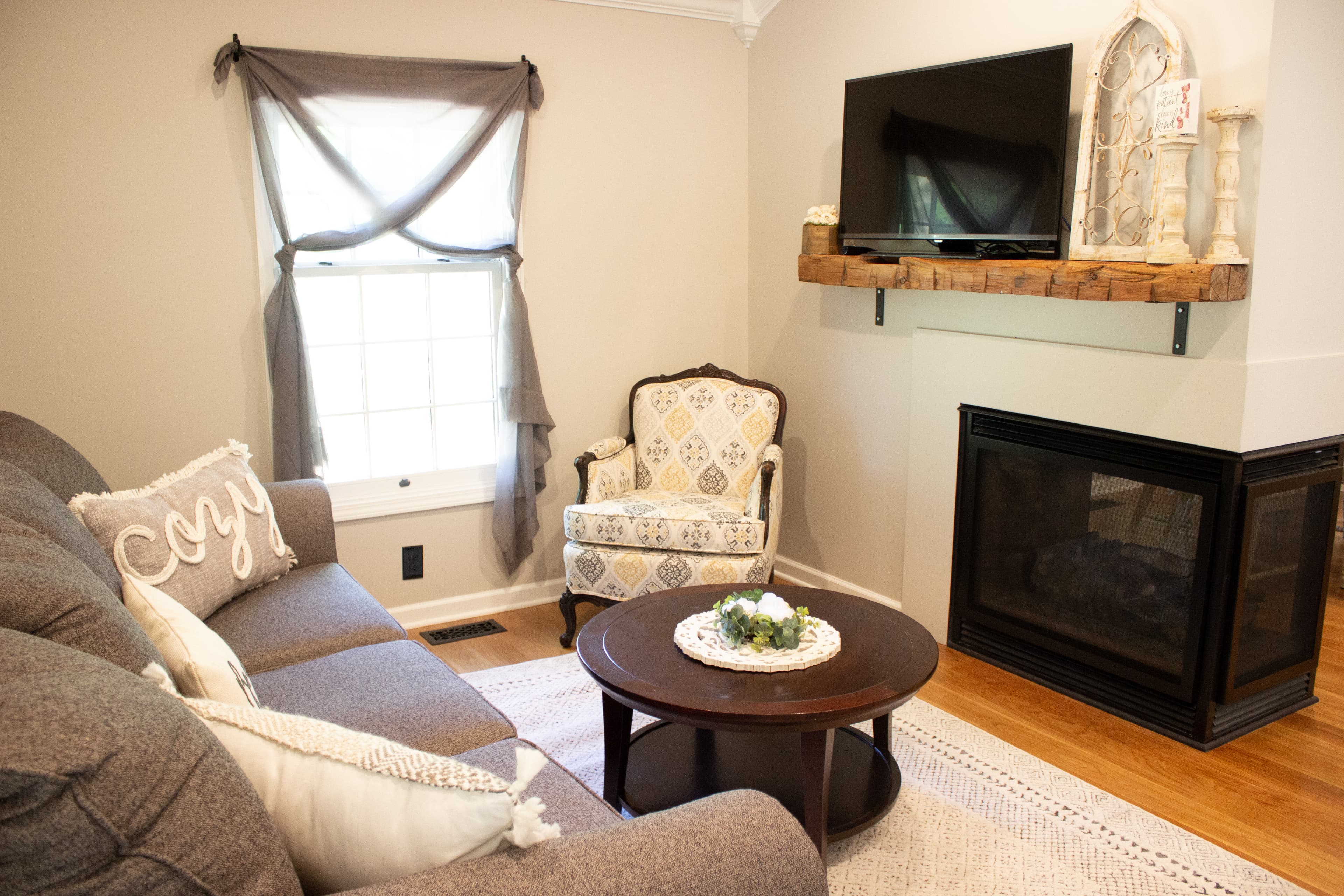 Cozy living room area at The August House featuring a corner fireplace with a wooden beam mantel and mounted TV, a grey sofa with accent pillows, and a patterned armchair.