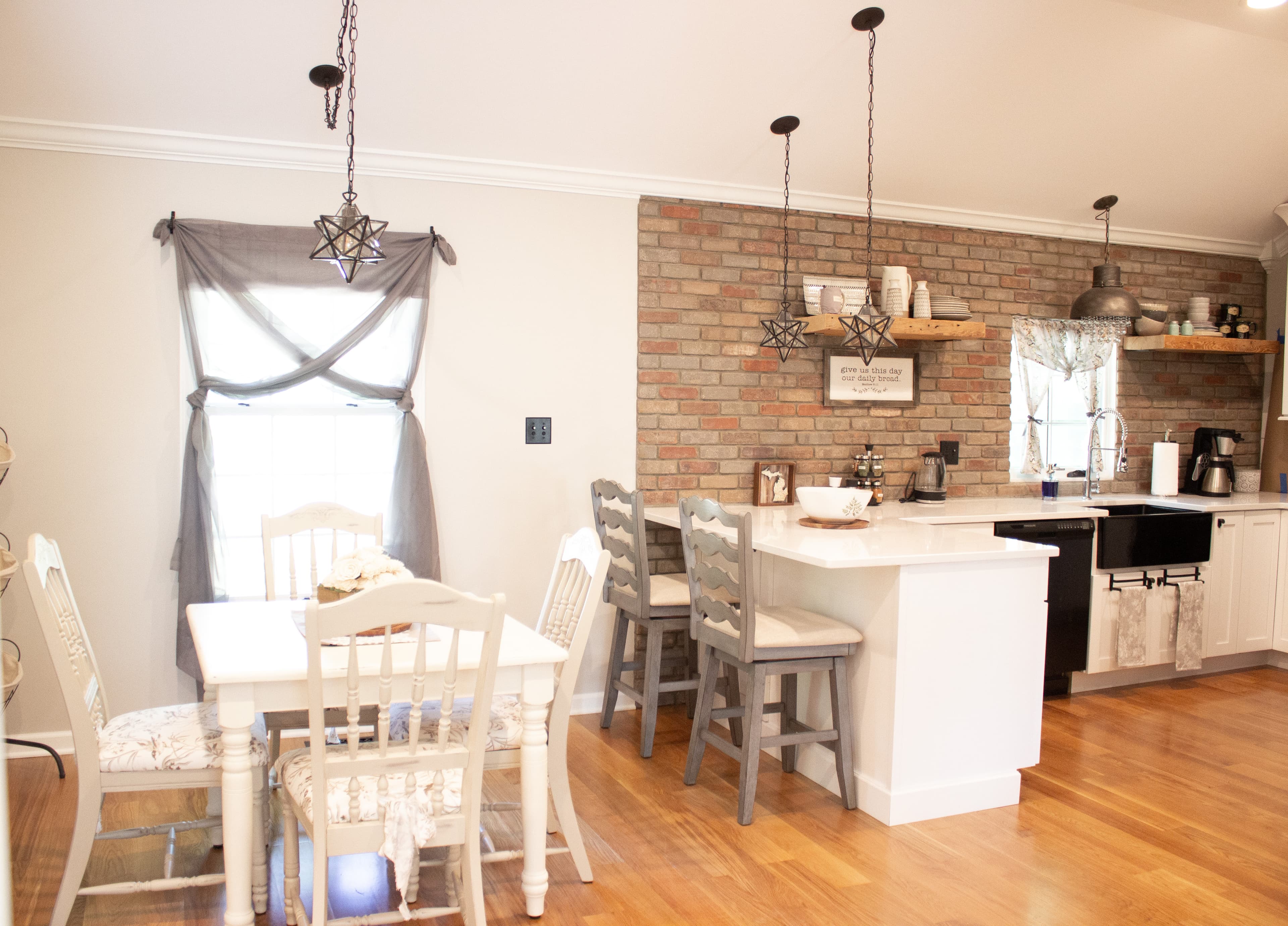 Open-concept farmhouse kitchen and dining area at The August House featuring a brick accent wall, white island with bar seating, and a dining table near a window with grey tie-up curtains.