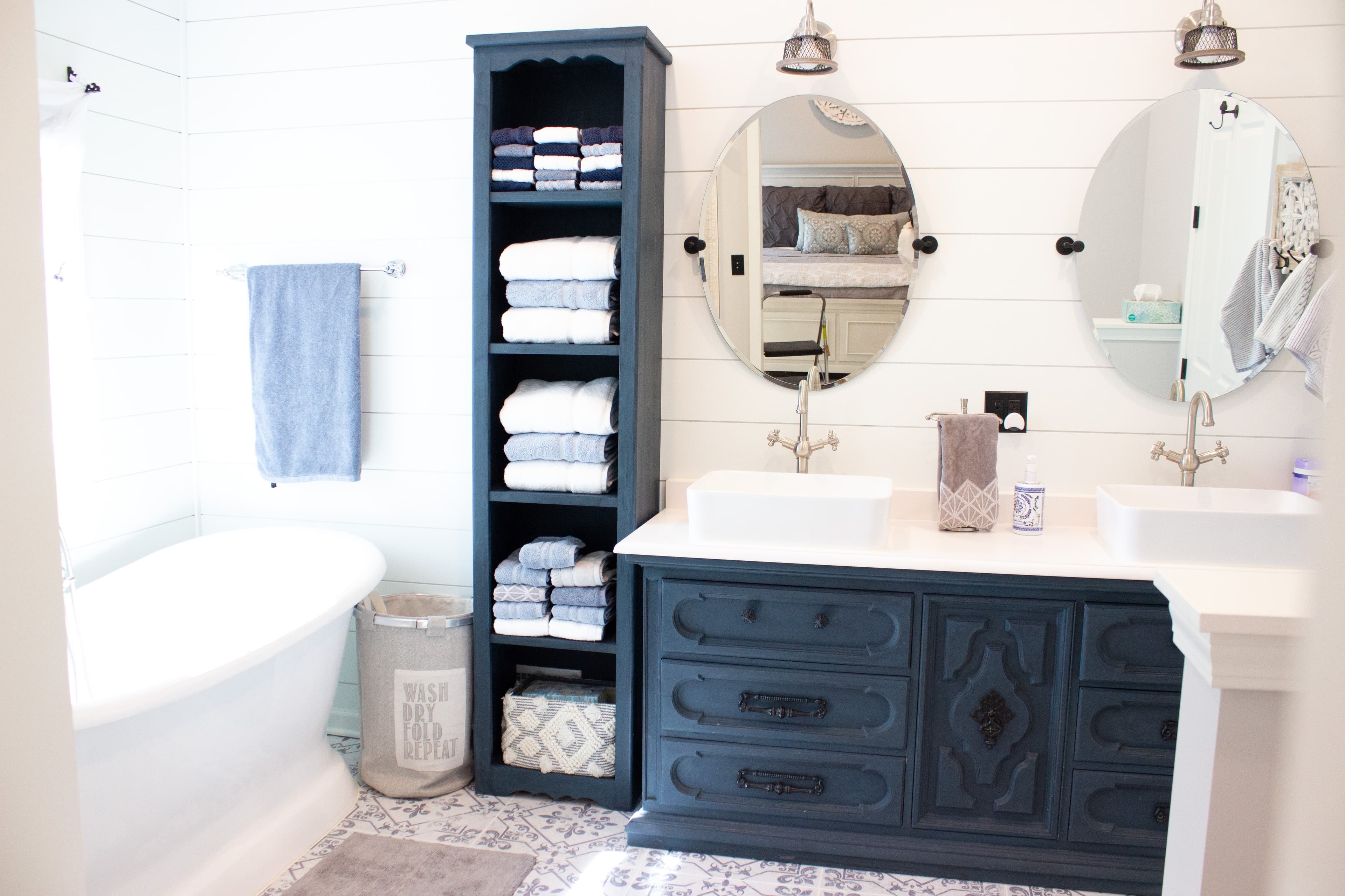 Spa-inspired farmhouse bathroom at The August House featuring a white freestanding soaking tub, navy blue double vanity with dual oval mirrors, and a tall linen tower against white shiplap walls.