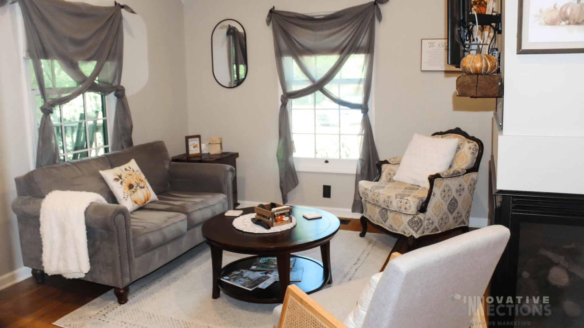 Cozy living room featuring a gray sofa, patterned chair, round coffee table, and large windows with flowing curtains.