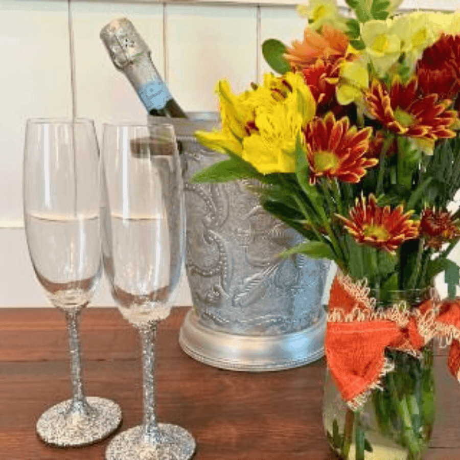 Two sparkling glasses beside a silver ice bucket holding a champagne bottle and a colorful flower bouquet.