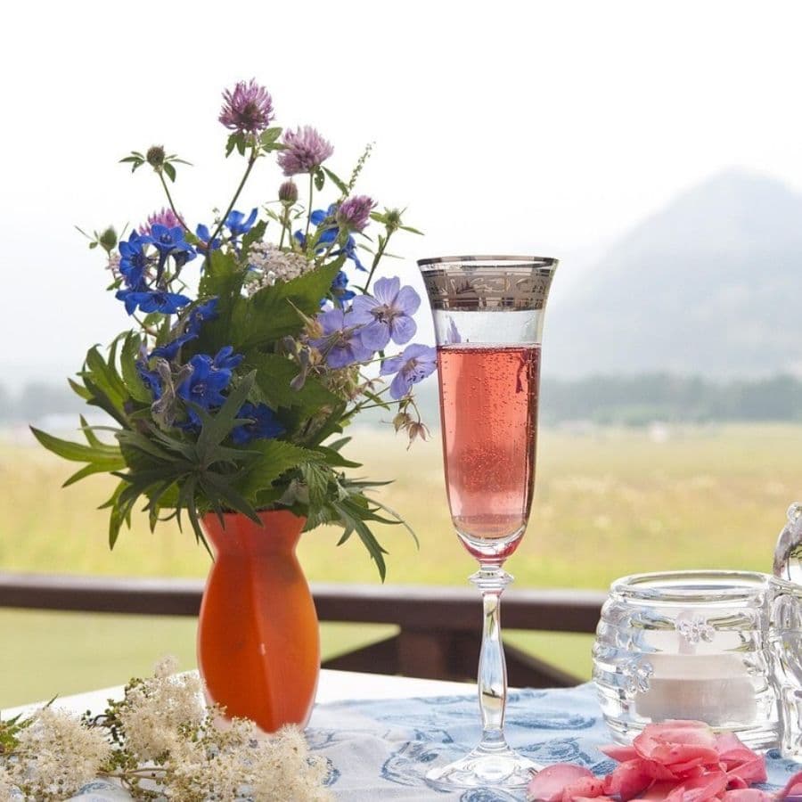 A glass of pink sparkling drink next to a vibrant bouquet of blue and purple flowers on a table with a scenic background.