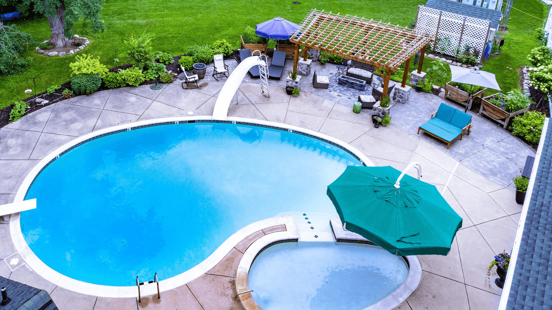 Aerial view of a landscaped backyard featuring an oval swimming pool, hot tub, and seating area with umbrellas.