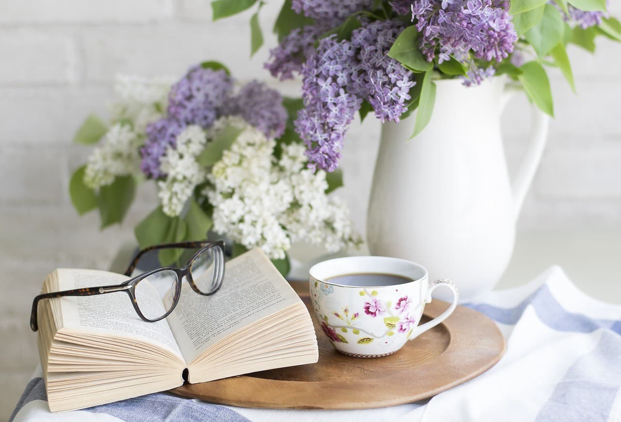 A cozy scene featuring an open book, glasses, a floral teacup, and a bouquet of lilacs. A cozy scene featuring an open book, glasses, a floral teacup, and a bouquet of lilacs.