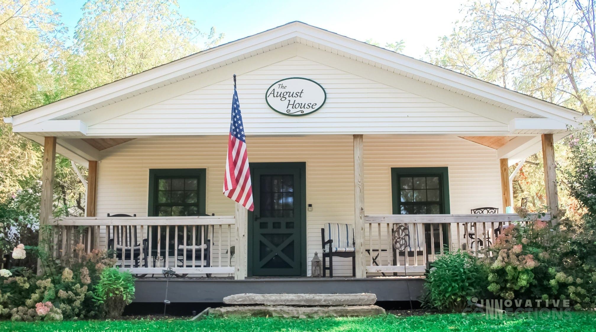 A white cottage with green trim and a front porch, featuring an American flag and a sign reading "The August House."
