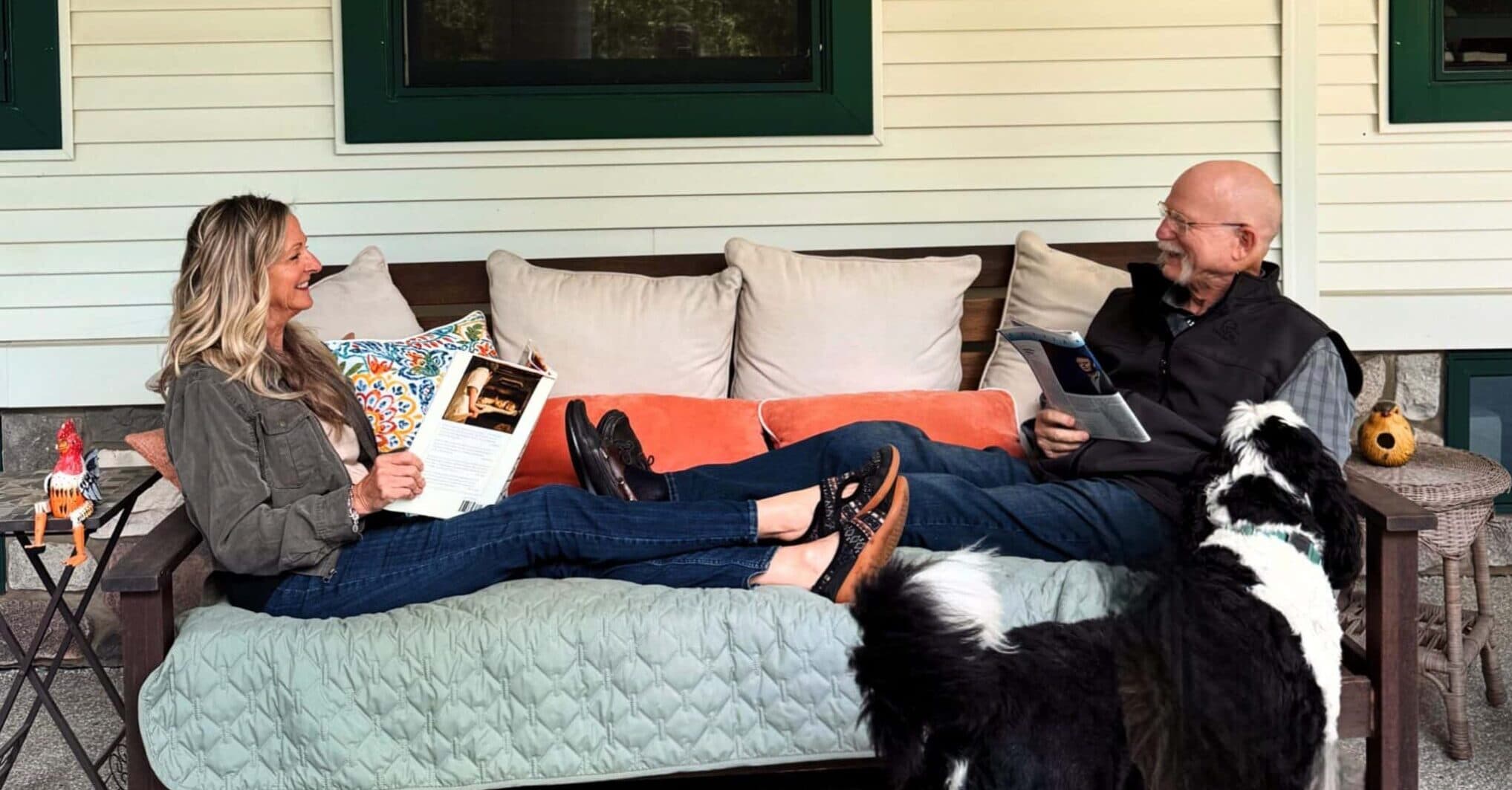 A woman and a man sit on a couch, enjoying books and an accompanying dog on a porch.