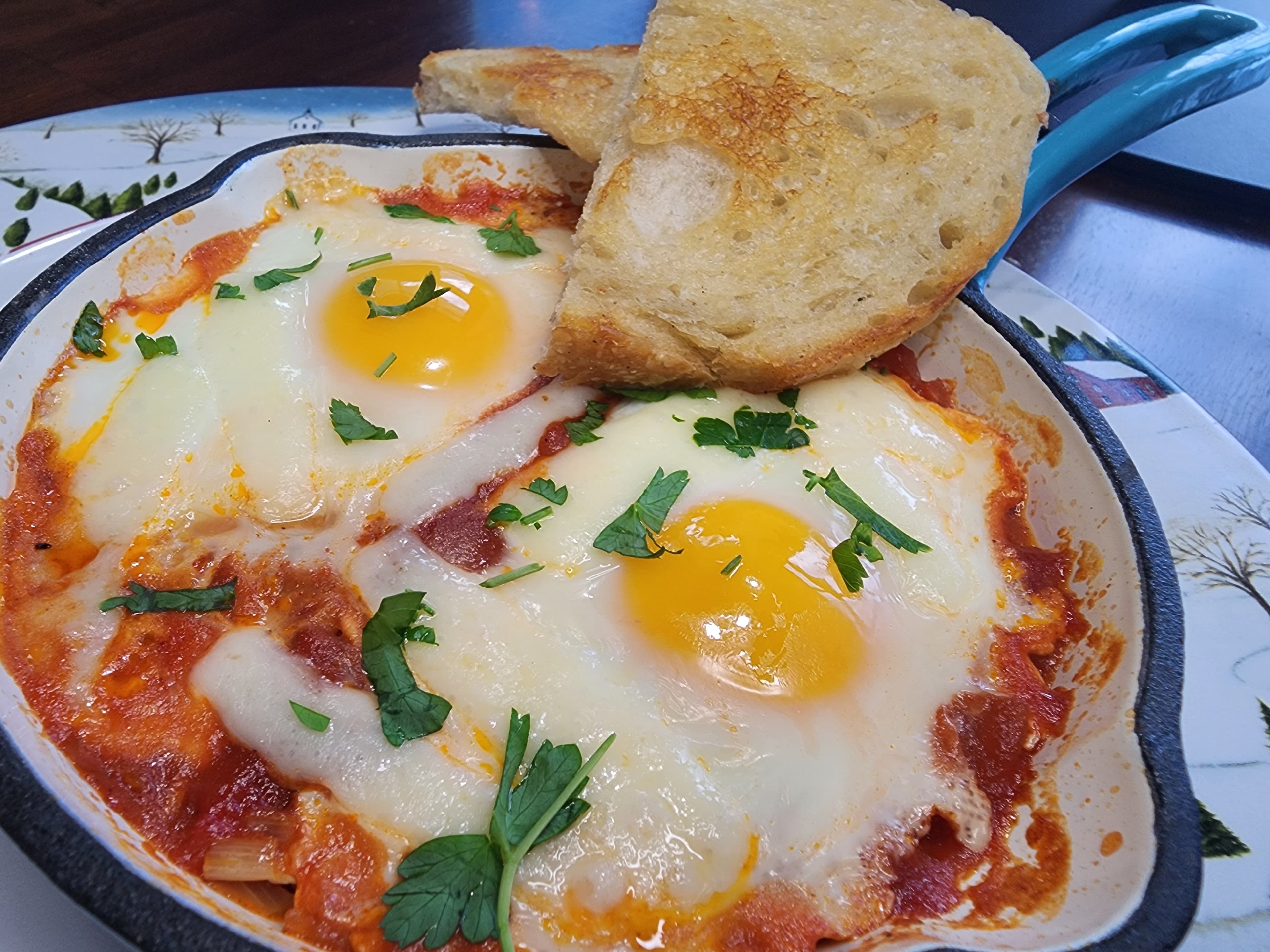 A skillet of baked eggs in tomato sauce, garnished with parsley and served with a slice of toasted bread.