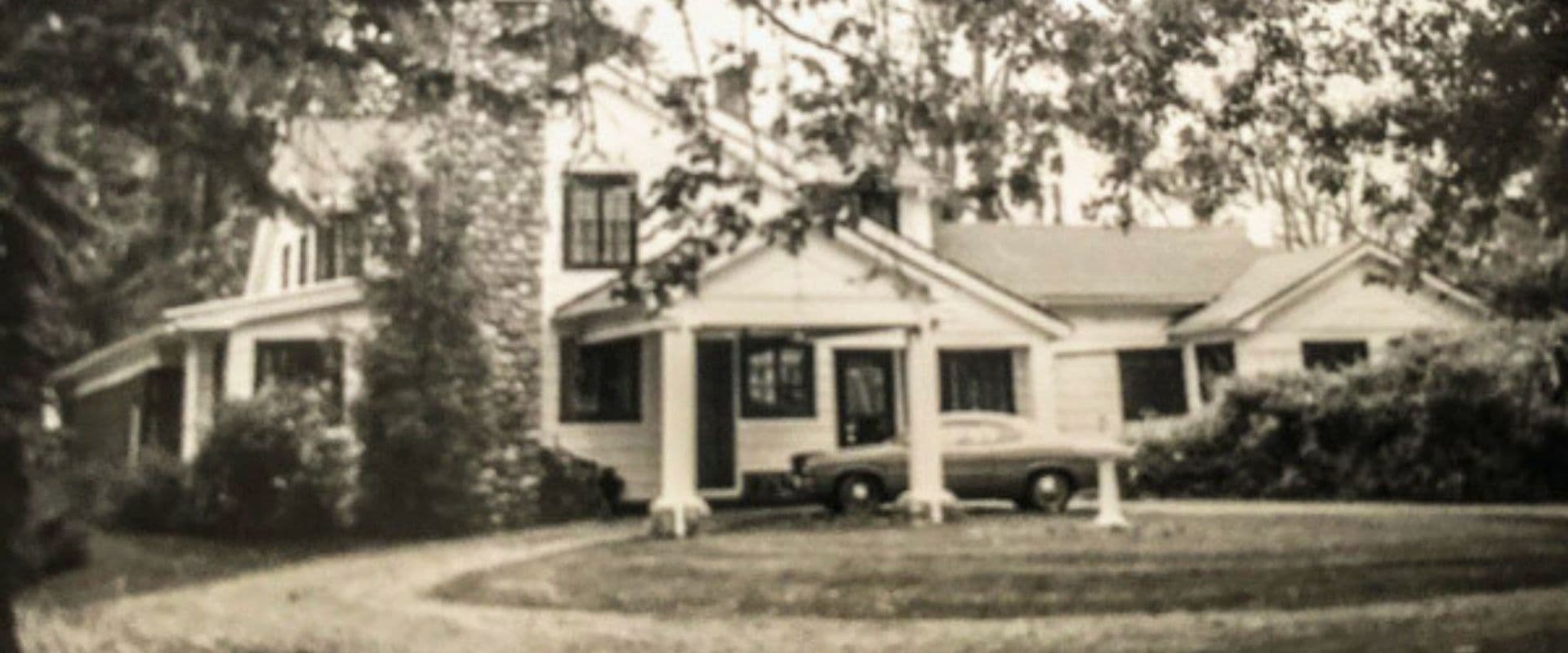 A black-and-white photo of a large, vintage house surrounded by trees and a lawn, featuring a car parked in the front.