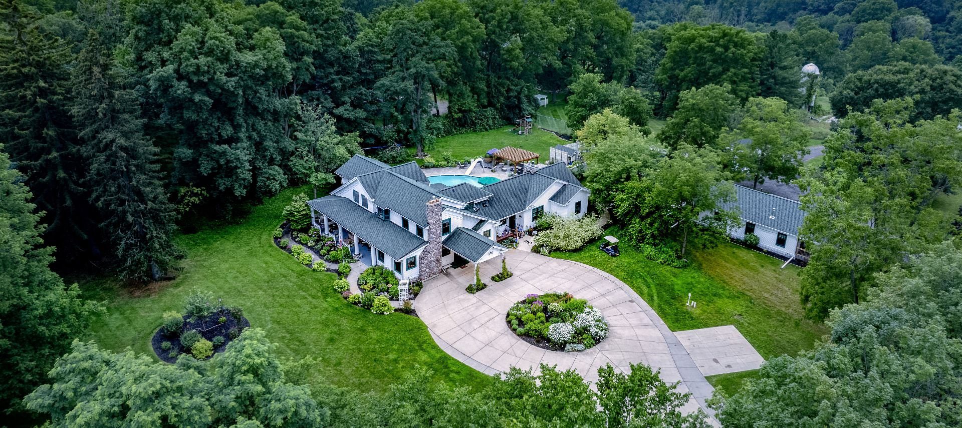 Aerial view of the property painted white with white and teal trim, stone fireplace, large driveway, and surrounded by lush trees, bushes, grass, and flowers