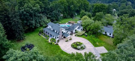 Aerial view of the property painted white with white and teal trim, stone fireplace, large driveway, and surrounded by lush trees, bushes, grass, and flowers