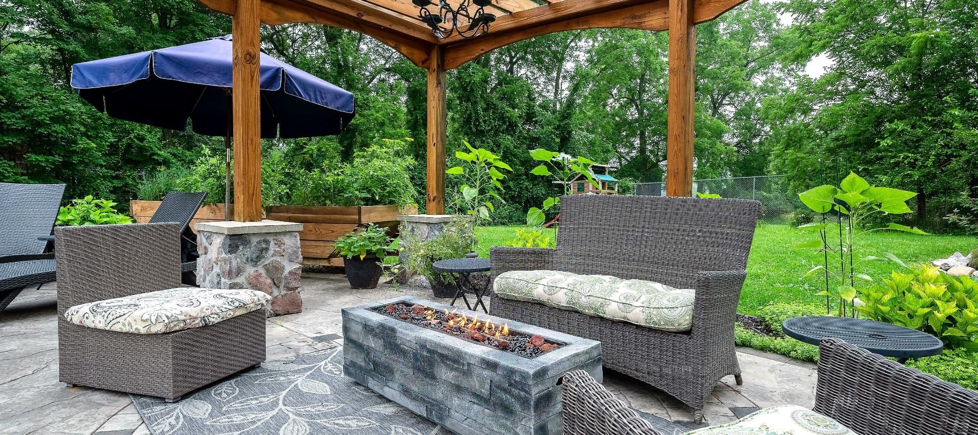 Concrete patio with gray wicker patio furniture, fire pit, and wooden pergola surrounded by green trees and grass