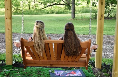 Two women with backs to the camera sitting on a wooden bench swing looking out at the green grass and trees relaxing