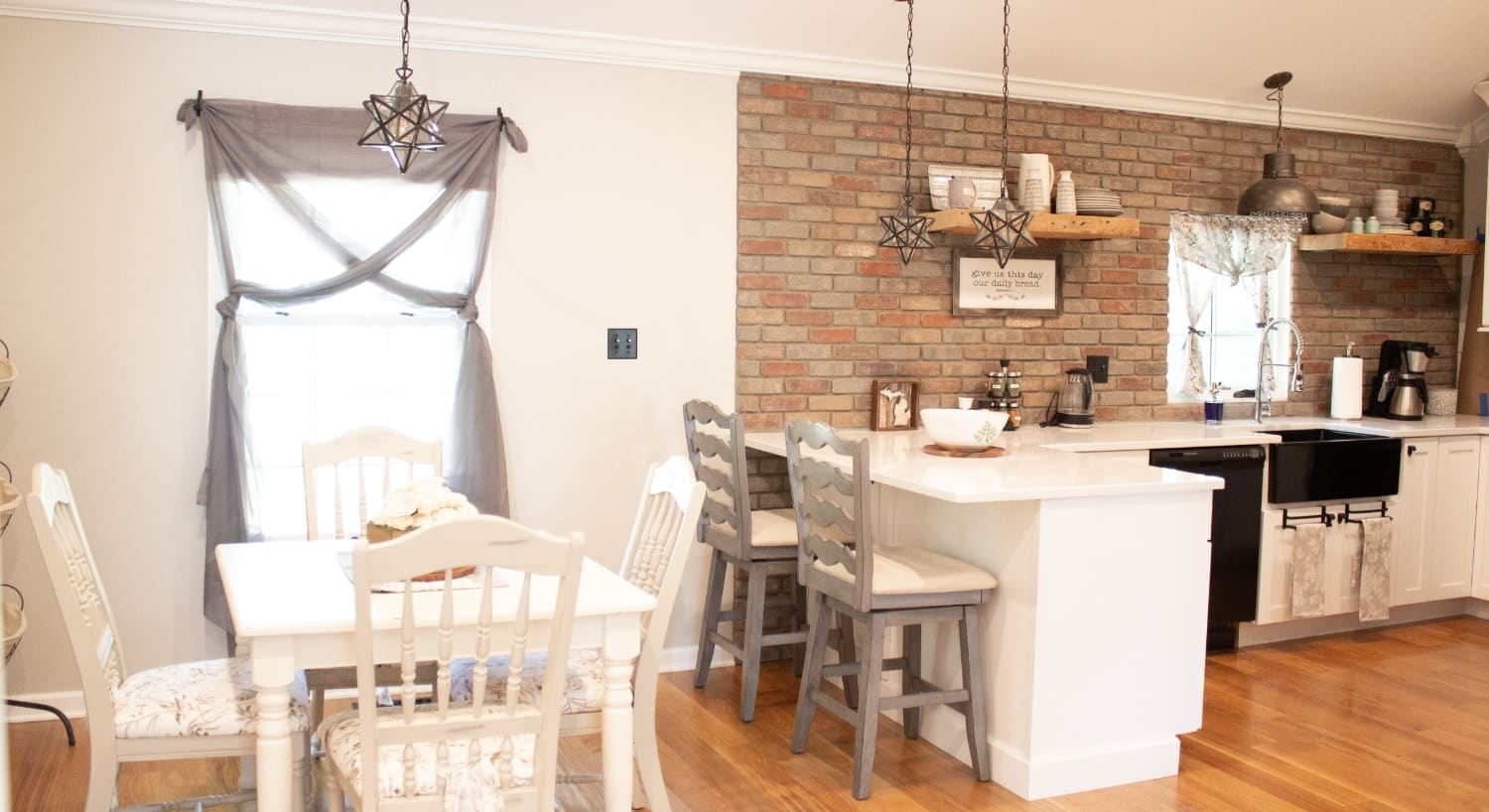 Kitchen and dining area with white walls, a brick accent wall, hardwood flooring, white cabinets, black appliances, and hanging star shaped lights