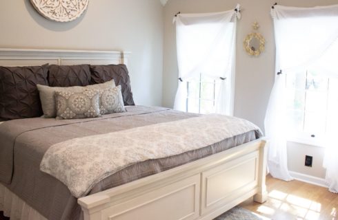 Bedroom with white walls, hardwood flooring, white wooden bed, light gray bedding, and two windows with wispy white curtains