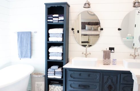 Bathroom with white panel walls, white claw foot tub, navy vanity, double white sinks, and double oval mirrors