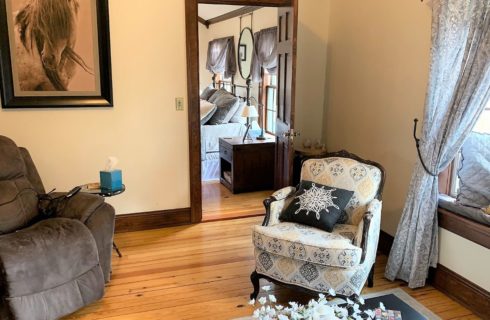 Room with white walls, hardwood flooring, oversized gray upholstered recliner, and multicolored upholstered armchair with a view into the bedroom