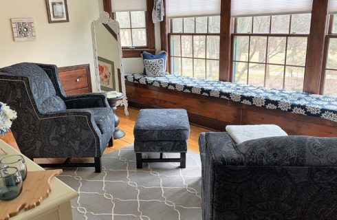 Two paisley blue upholstered armchairs with ottomans in a room with white walls, hardwood flooring, and long window seat next to multiple windows