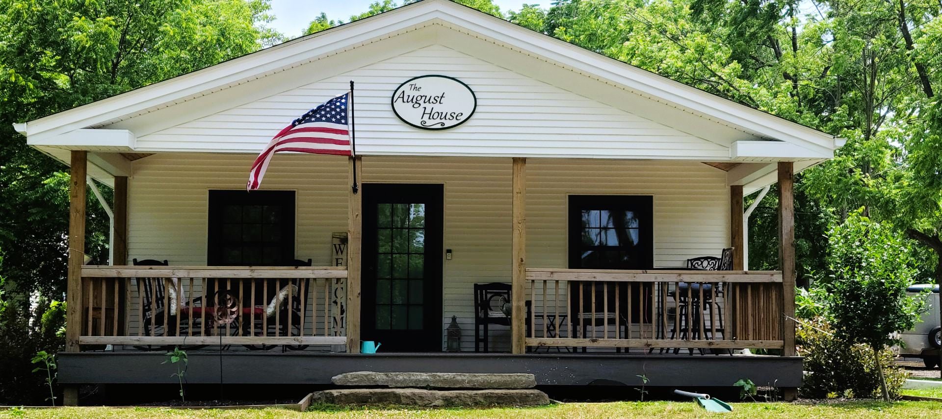 Exterior view of the August House, painted white with dark trim, front porch, and surrounded by green trees and grass