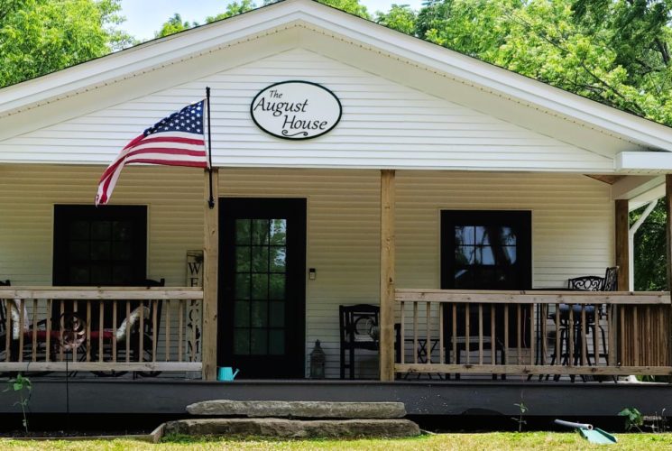 Exterior view of the August House, painted white with dark trim, front porch, and surrounded by green trees and grass