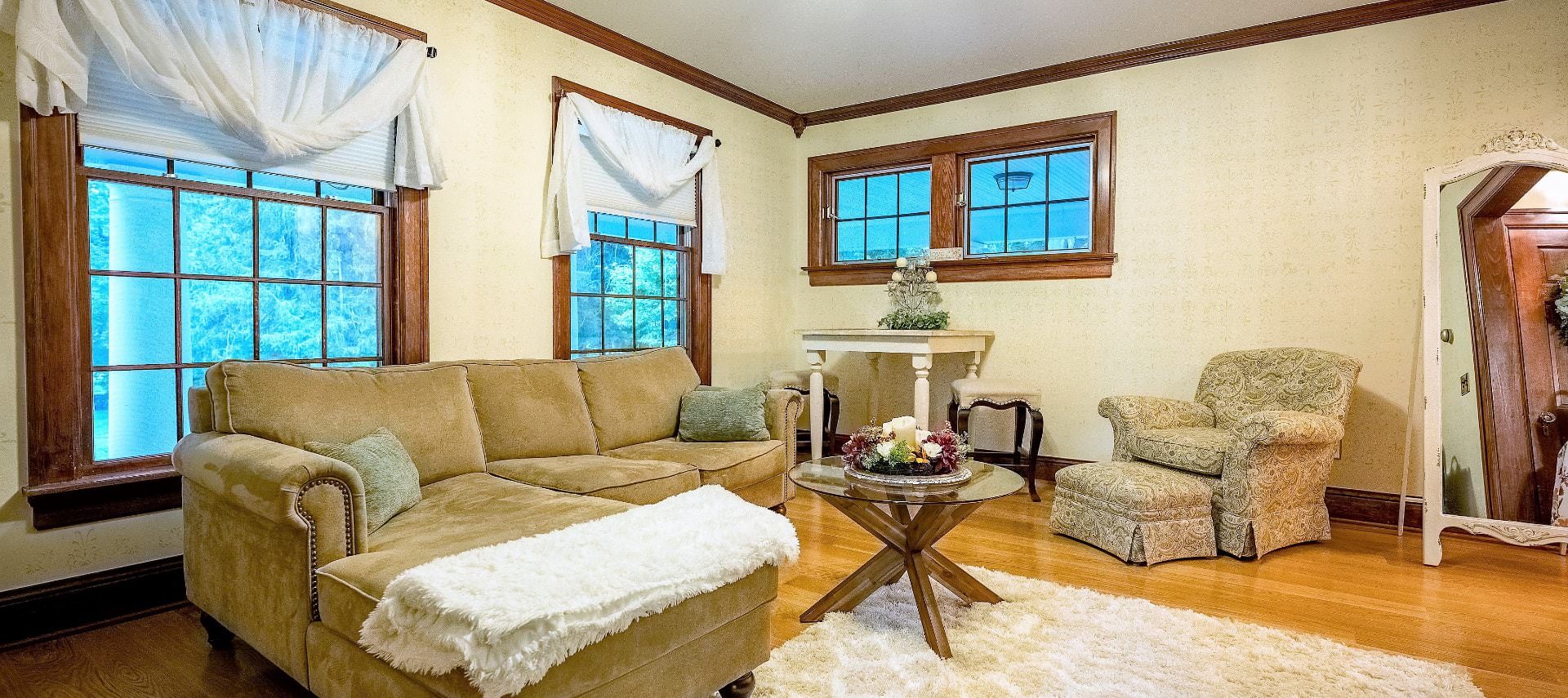 Large living room with white walls, hardwood flooring, light tan upholstered couch with chaise, light colored upholstered armchair, and multiple windows