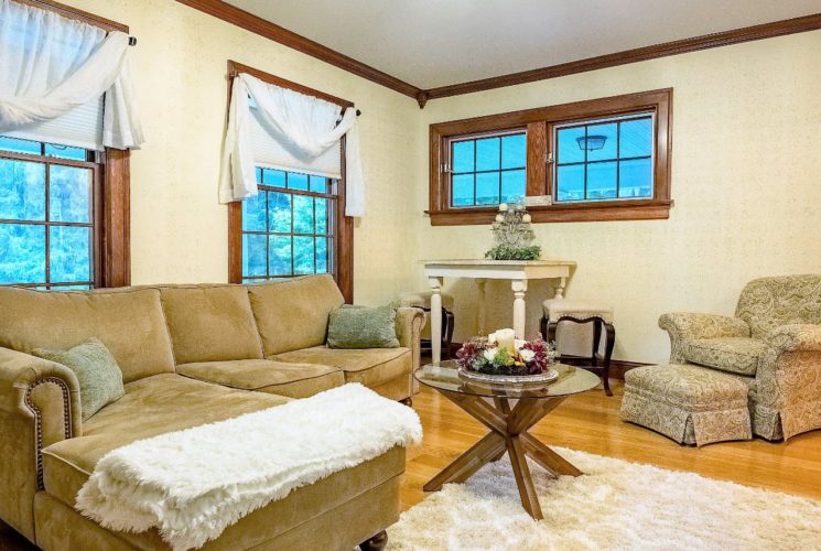 Large living room with white walls, hardwood flooring, light tan upholstered couch with chaise, light colored upholstered armchair, and multiple windows