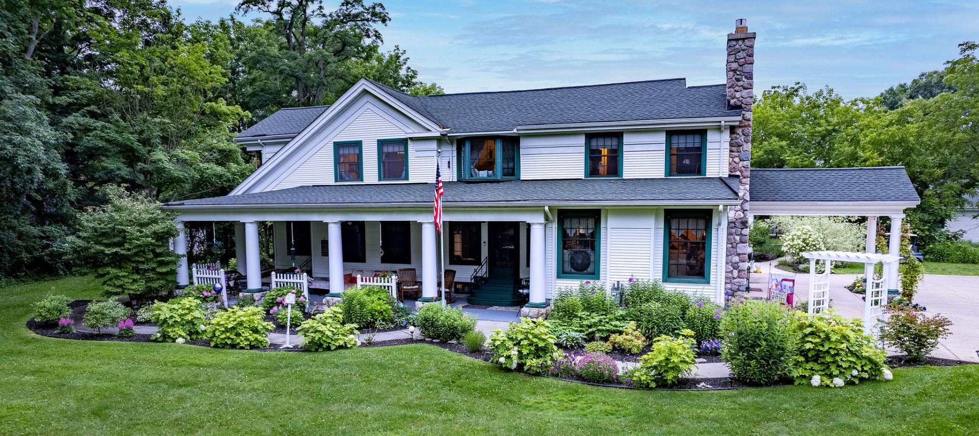 Exterior view of the property painted white with white and teal trim, stone fireplace, large driveway, and surrounded by lush trees, bushes, grass, and flowers