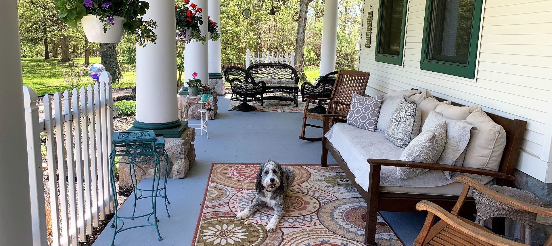 Front porch with dark wicker and dark wooden patio furniture and rocking chairs, area rugs, and gray and white dog laying on a rug