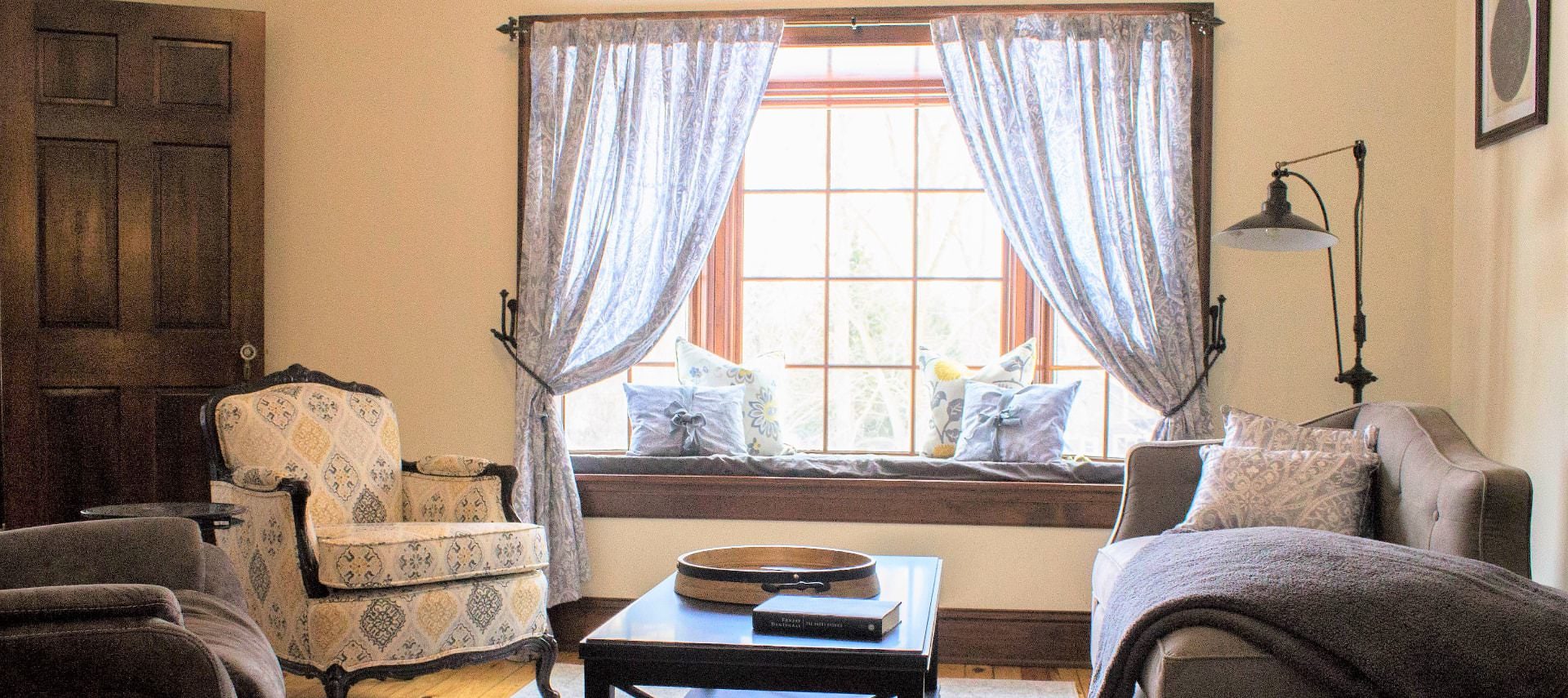 Room with a multicolored upholstered armchair, gray upholstered chair and loveseat, and dark wooden coffee table next to a bay window