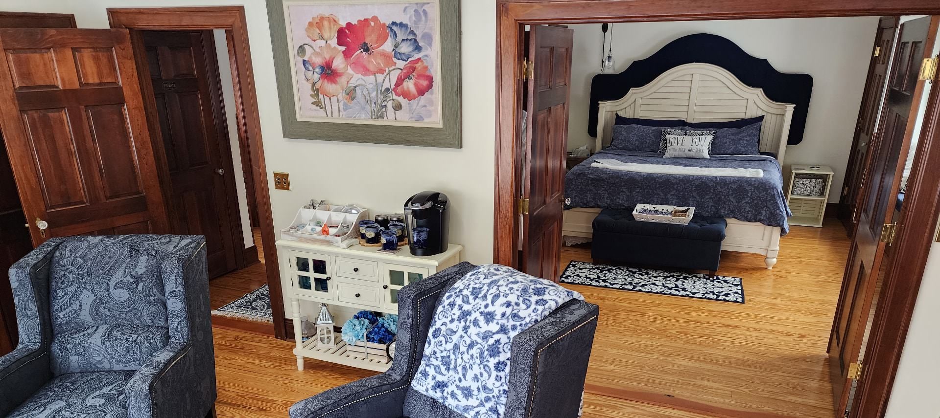 Room with two paisley blue upholstered armchairs, white walls, hardwood flooring, and a view into the bedroom