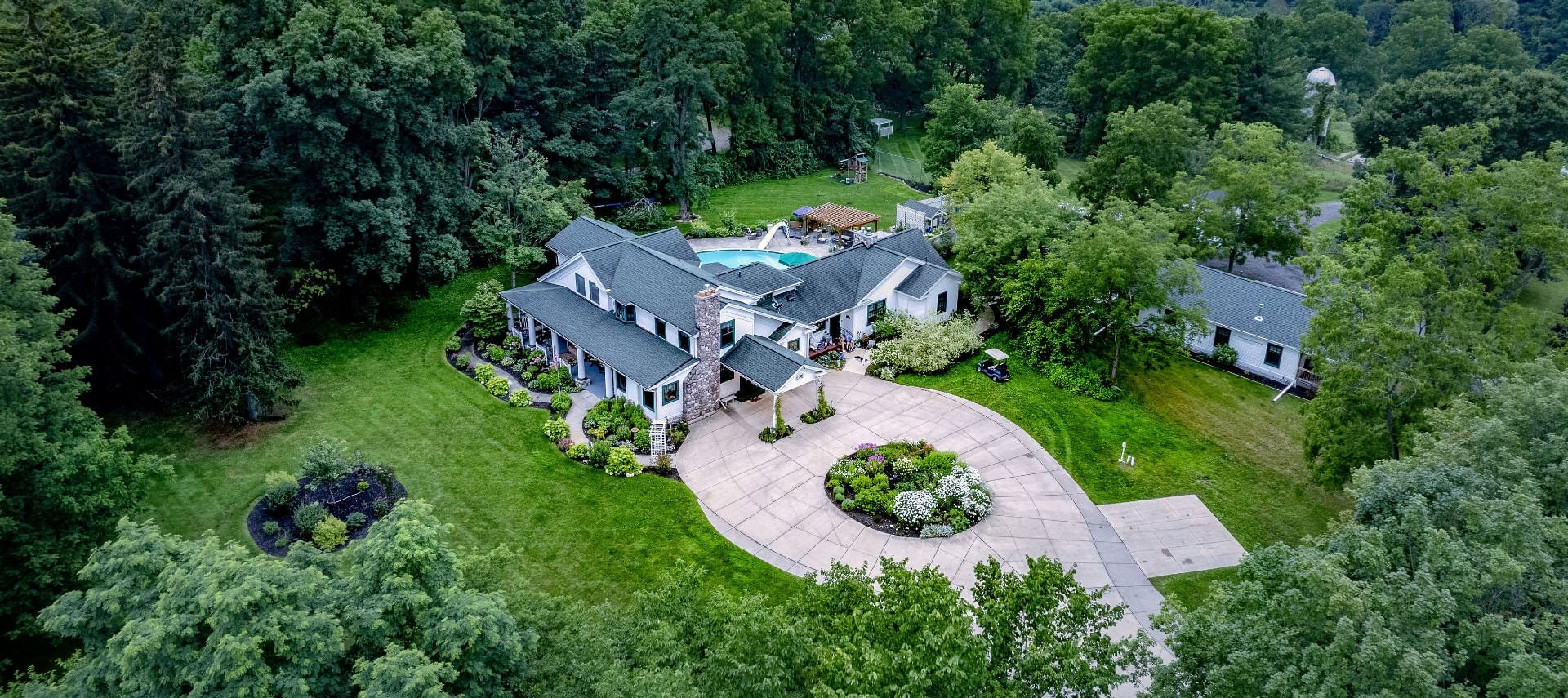 Aerial view of the property painted white with white and teal trim, stone fireplace, large driveway, and surrounded by lush trees, bushes, grass, and flowers