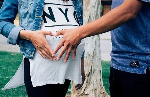 Close up view of a pregnant woman wearing a long white shirt with her hand and a man's hand forming the shape of a heart near her belly