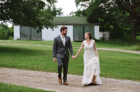Groom with gray suit walking and holding hand of bride in white dress surrounded by green trees and grass
