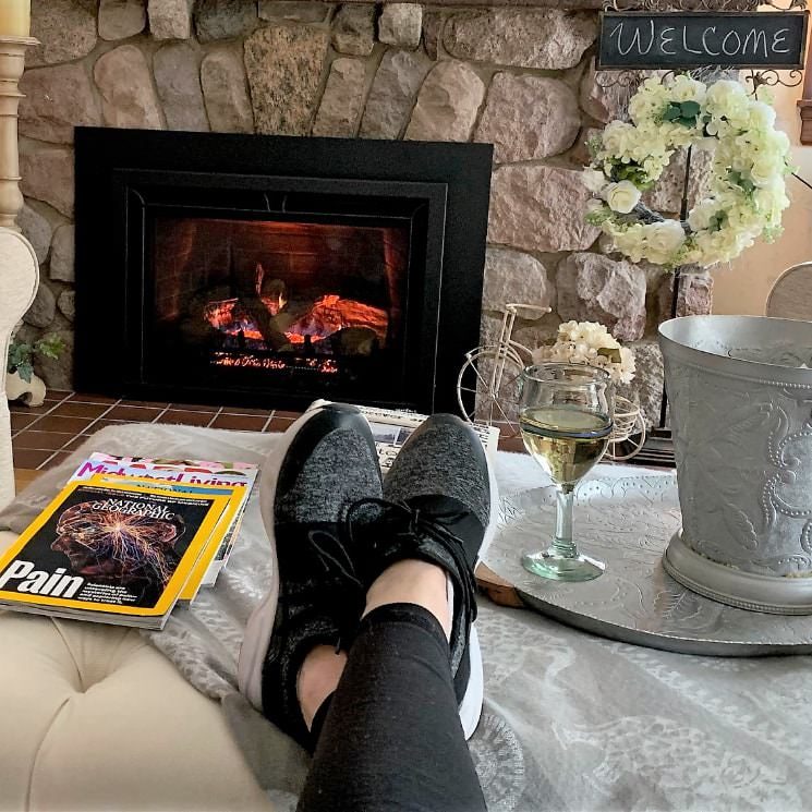 Person's black and gray shoes resting on a light ottoman next to magazines and tray with a glass of white wine with a stone fireplace in the background