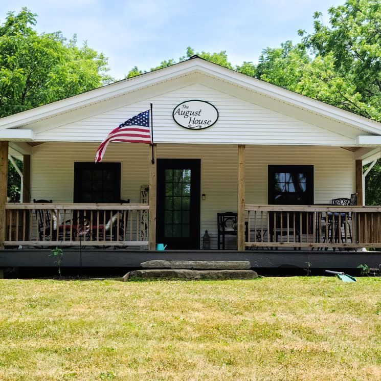 Exterior view of the August House painted white with dark trim and front porch surrounded by green trees and grass