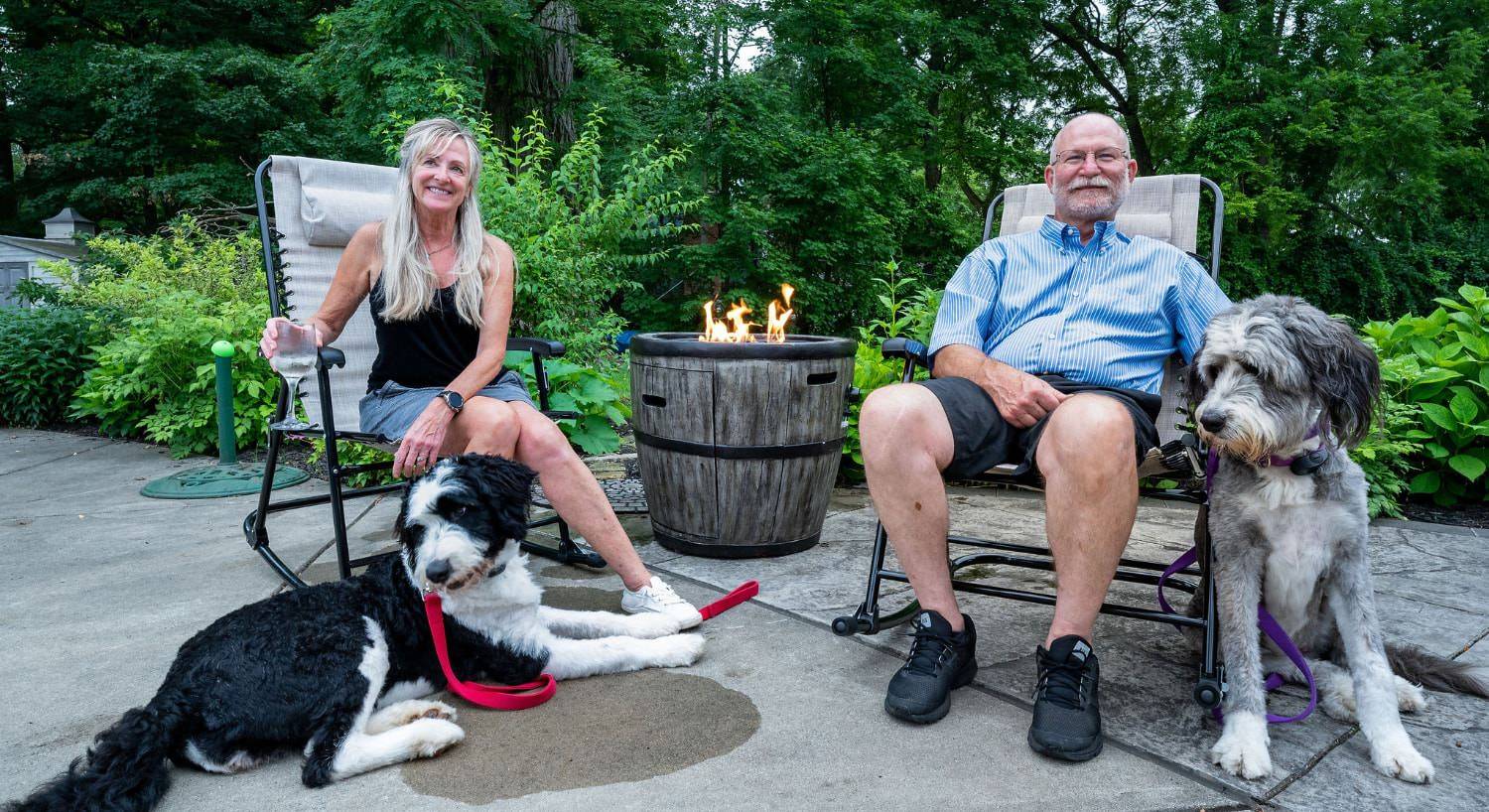 Man and woman sitting in chairs on concrete patio next to a fire pit with a large black and white dog and a large gray and white dog with greenery in the background