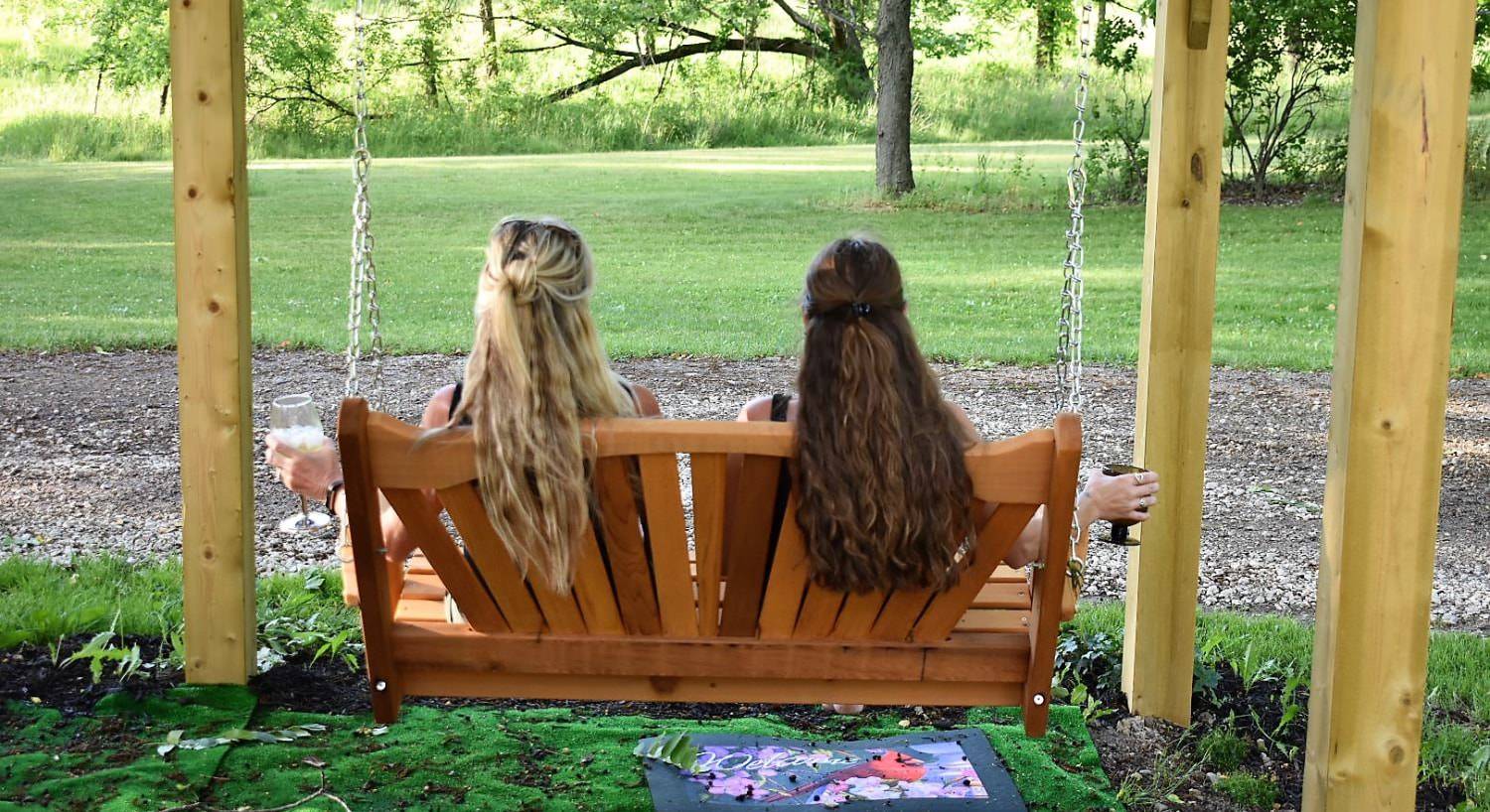 Two women with backs to the camera sitting on a wooden bench swing looking out at the green grass and trees